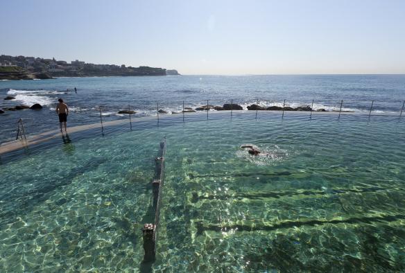 Une personne nageant dans l'eau étincelante des Bronte Baths et une autre personne debout au bord de la piscine, surplombant l'océan à Bronte Beach, Sydney, Nouvelle-Galles du Sud © Andrew Gregory/Destination NSW
