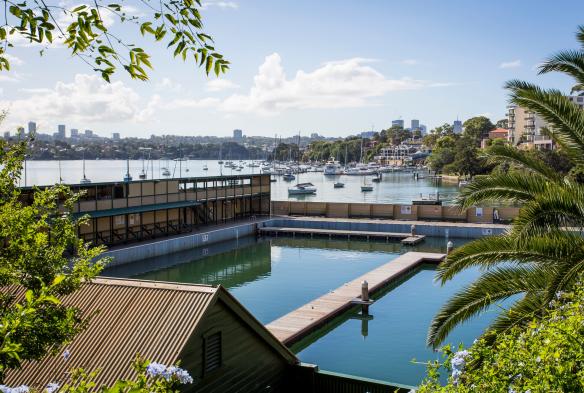 Vue à travers les arbres des piscines des Dawn Fraser Baths, avec des bateaux et des bâtiments en front de mer en arrière-plan à Balmain, Sydney, Nouvelle-Galles du Sud © Destination NSW