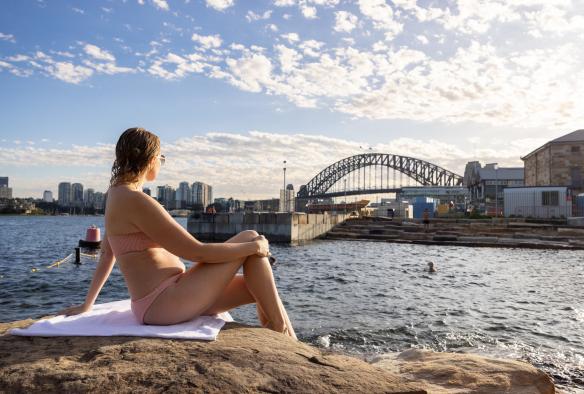 Une femme assise sur un rocher de grès regardant au-delà de Marrinawi Cove, en direction du Sydney Harbour Bridge, pendant que des personnes nagent en contre-bas, Barangaroo Reserve, Sydney, Nouvelle-Galles du Sud © Images for Business