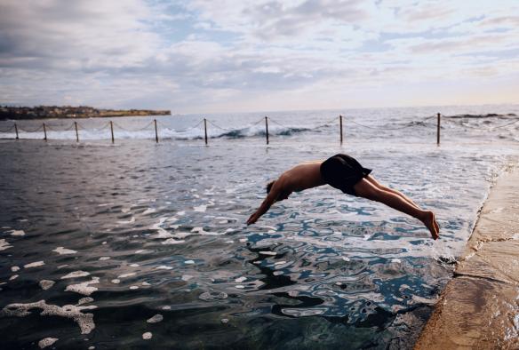 Un homme plongeant dans l'eau des Wylie's Baths, avec des vagues déferlant dans l'océan en arrière-plan à Coogee, Nouvelle-Galles du Sud © Destination NSW