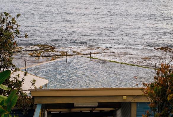 Vue sur l'entrée des Wylie's Baths et de leur eau paisible dans laquelle une personne nage au premier plan, avec l'océan en arrière-plan, Coogee, Nouvelle-Galles du Sud © Destination NSW