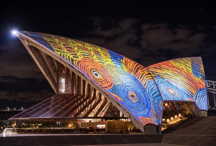 Layar Sydney Opera House menjadi terang dengan proyeksi cahaya yang mengilustrasikan desain Penduduk Asli berwarna biru, oranye, dan kuning, Sydney, New South Wales © Daniel Boud