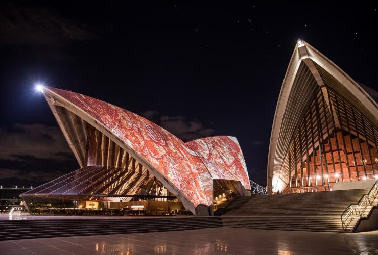 Langit malam nan gelap dengan layar bercahaya terang di Sydney Opera House yang mengilustrasikan seni Penduduk Asli berwarna merah, Sydney, New South Wales © Daniel Boud