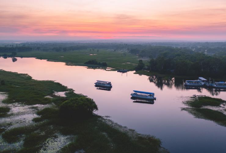 Pemandangan dari udara di atas perahu-perahu yang berlayar di sepanjang sungai tenang yang dikelilingi oleh tanaman hijau, memantulkan warna merah muda pastel dan jingga di langit saat matahari terbenam di kolam billabong Yellow Water, Kakadu National Park, Northern Territory © Tourism NT/Salty Wings