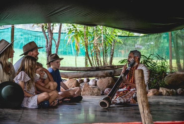 Keluarga duduk di lantai di samping pemandu Aborigin yang mengenakan cat tubuh dan memegang didgeridoo di Rainforestation Nature Park, Kuranda, Queensland © Tourism and Events Queensland