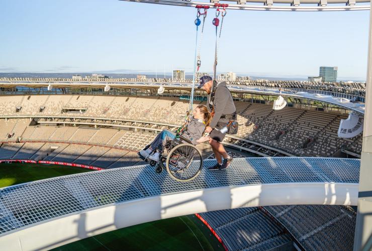 Seorang wanita yang duduk di kursi roda didorong menelusuri jalur tinggi sambil mengenakan tali pengaman di Ozone di Optus Stadium, Perth, Western Australia © Tourism Western Australia
