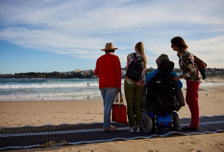 Seorang pria di kursi roda duduk di pantai di atas tikar dengan keluarganya, Australia In Style, Bondi Beach, Sydney, New South Wales © Destination NSW