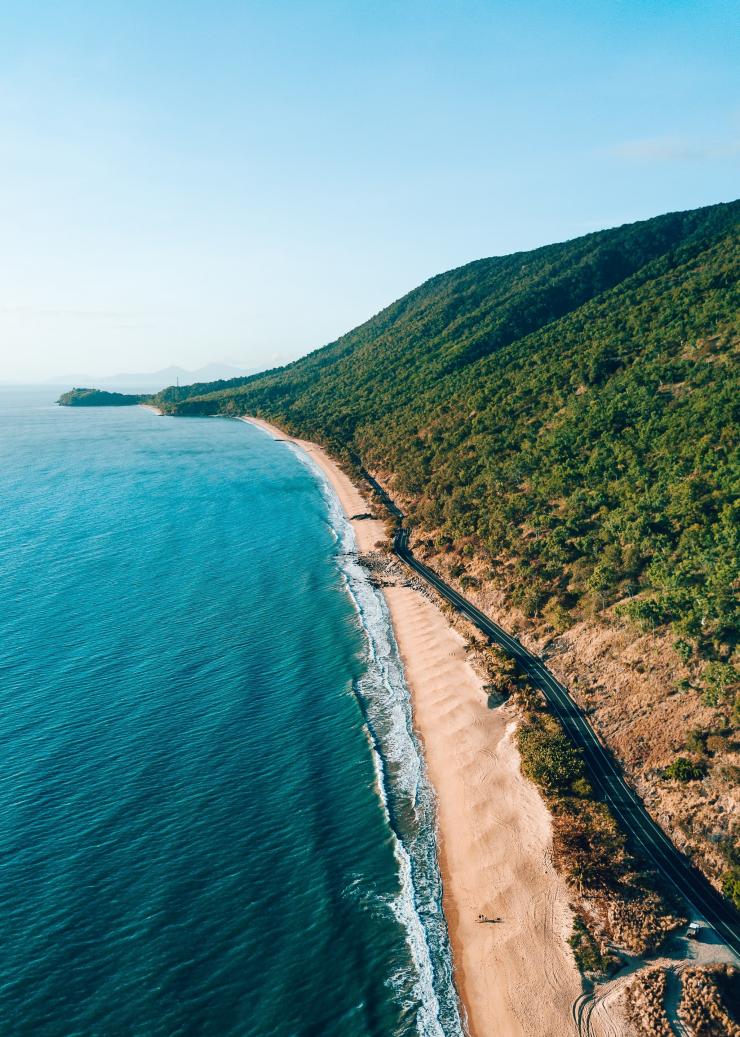 Pemandangan dari udara jalan yang membentang di sepanjang pantai berpasir keemasan dengan gunung tinggi yang ditutupi hutan hujan di sisi lainnya di sepanjang Great Barrier Reef Drive, Tropical North Queensland, Queensland © Tourism and Events Queensland
