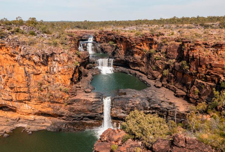 Dua orang berdiri di dekat air terjun besar yang bertingkat-tingkat di kolam di Mitchell Falls, East Kimberley, Western Australia © Tourism Australia