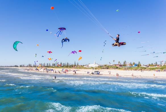 Dua orang melakukan wakeboarding di udara di tengah langit yang dipenuhi layang-layang warna-warni, diterbangkan oleh orang-orang yang memadati pantai berpasir putih di Semaphore Beach, Semaphore, South Australia © Michael Waterhouse Photography