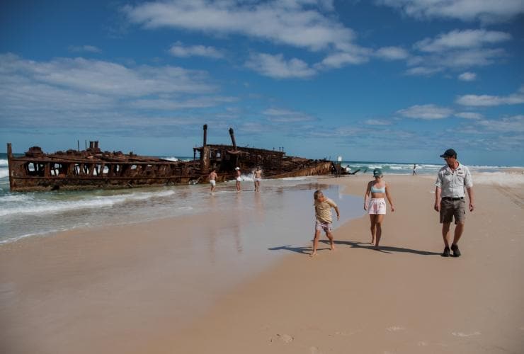 Dua anak berjalan bersama pemandu tur di pantai dengan sekelompok orang yang menjelajahi bangkai kapal di pantai di belakang mereka di dekat Kingfisher Bay Resort, K'gari, Queensland © Tourism and Events Queensland