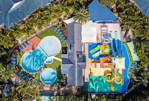 Pemandangan udara di atas akomodasi yang dikelilingi pepohonan dengan kolam renang dan taman air berwarna-warni di Turtle Beach Resort, Gold Coast, Queensland © Turtle Beach Resort