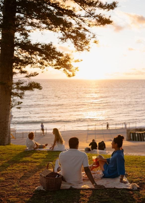 Sepasang pengunjung sedang berpiknik di bukit berumput dengan pemandangan samudra saat matahari terbenam, Cottesloe Beach, Perth, Western Australia © Tourism Australia