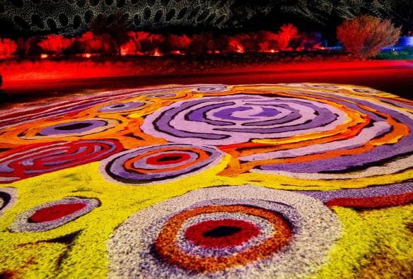 Dua orang berdiri di tengah kaleidoskop instalasi seni warna-warni yang dihias dengan foto burung-burung asli tempat ini di Parrtjima - A Festival in Light, Alice Springs, Northern Territory © Tourism NT/James Horan