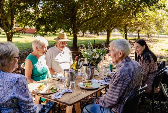 Sekelompok orang duduk di meja yang dikelilingi pohon-pohon macadamia di Macadamia Australia, Bundaberg, Queensland © Tourism Australia 