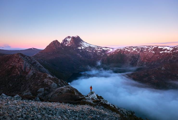 Seseorang berdiri di puncang gunung berbatu yang menghadap awan berketinggian rendah di atas lanskap pegunungan, Hansons Peak, Cradle Mountain, Tasmania © Daniel Tran