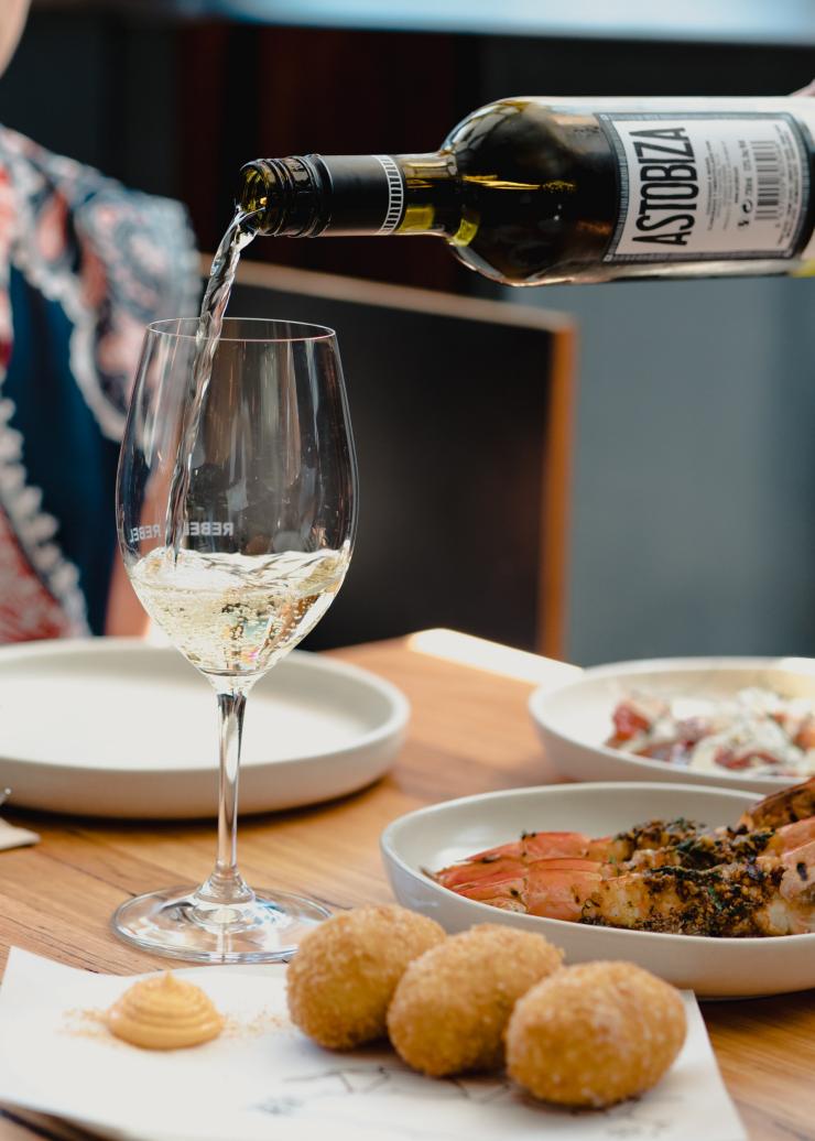 Seseorang menuangkan segelas white wine di atas meja yang menyajikan hidangan, Rebel Rebel, Canberra, Australian Capital Territory © Tourism Australia