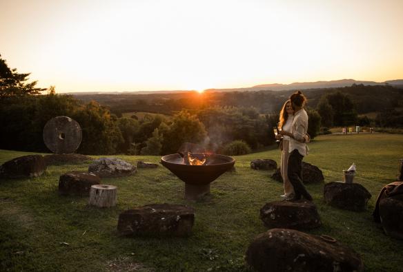 Sepasang kekasih berdiri di samping api unggun di bukit berumput saat matahari terbenam di Gaia Retreat & Spa, Byron Bay, New South Wales © Gaia Retreat & Spa