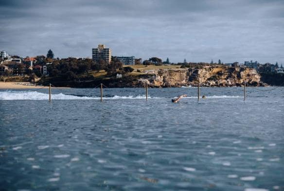 Foto close-up perairan biru Wylies Baths di latar depan dengan latar belakang orang yang berenang dan tanjung di belakangnya di Coogee, New South Wales © Destination NSW