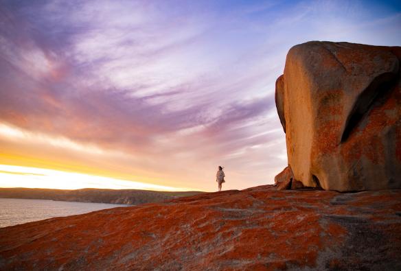 Seorang wanita berjalan di pinggir tebing yang tertutup lumut berwarna oranye yang menghadap ke samudra saat matahari terbit dengan Remarkable Rocks di sampingnya, Kangaroo Island, South Australia © Tourism Australia