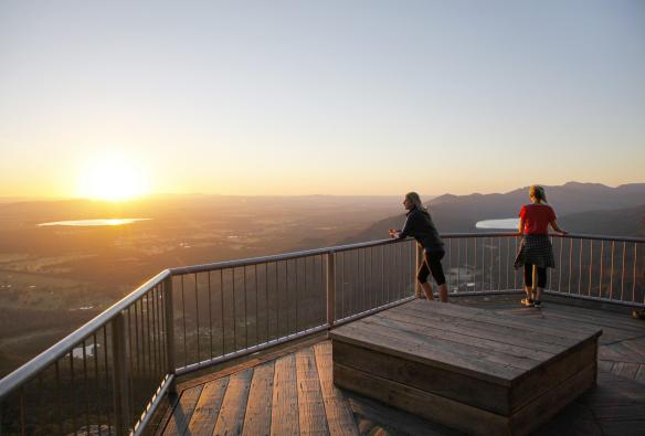 Dua orang berdiri di Boroka Lookout yang menghadap ke padang semak, jalur air, dan pegunungan saat matahari terbit, Grampians National Park, Victoria © Visit Victoria
