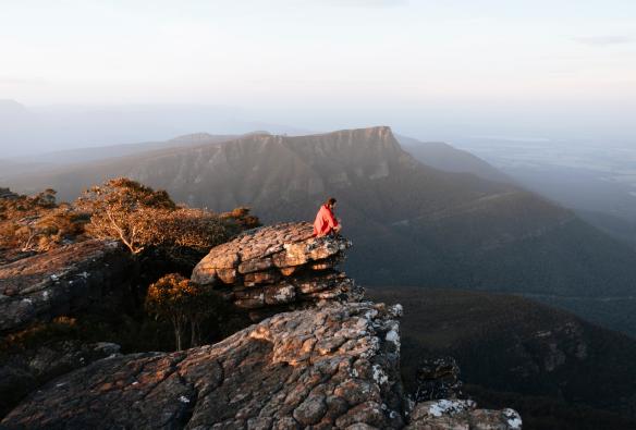 Seseorang duduk di tepi batu di puncak Mount William yang mengarah ke padang semak, Grampians National Park, Victoria © Ain Raadik Photography