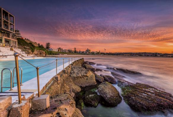 Perairan Bondi Icebergs yang berwarna biru cerah dengan langit pelangi dan ombak yang dengan lembut menerpa bebatuan di sekitarnya saat matahari terbit, Bondi Beach, New South Wales © Destination NSW