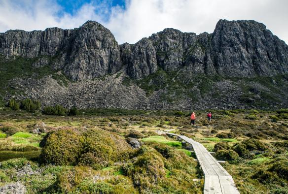 Dua penjelajah berjalan di jalur kecil yang terhampar di atas berbagai tumbuhan laut mengarah ke formasi batu yang menjulang di depan mereka di Walls of Jerusalem National Park, Tasmanian Wilderness World Heritage Area, Tasmania © O&M St John Photography