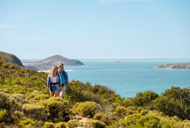Dua orang menjelajahi padang semak yang menghadap ke samudra biru di sepanjang Wild South Coast Way, Heysen Trail, Fleurieu Peninsula, South Australia © heidi who photos