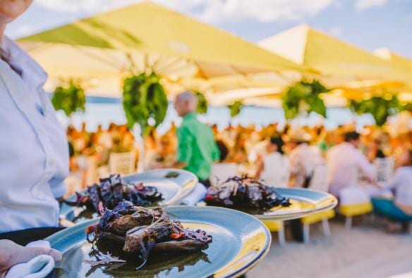 Un cameriere che porta pietanze impiattate con cura a un gruppo di persone che mangiano all'aperto durante il pranzo "Stranded at qualia" di Paspaley, Hamilton Island, Queensland © Tourism Australia 