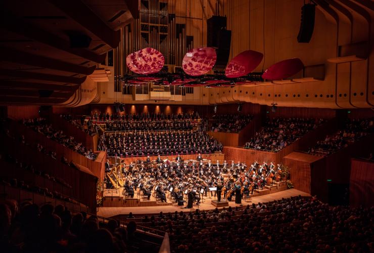 Foto panoramica della Sydney Symphony Orchestra disposta a semicerchio sul palco mentre esegue un brano davanti a un vasto pubblico di persone sedute presso la Sydney Opera House, Sydney, New South Wales © Daniel Boud