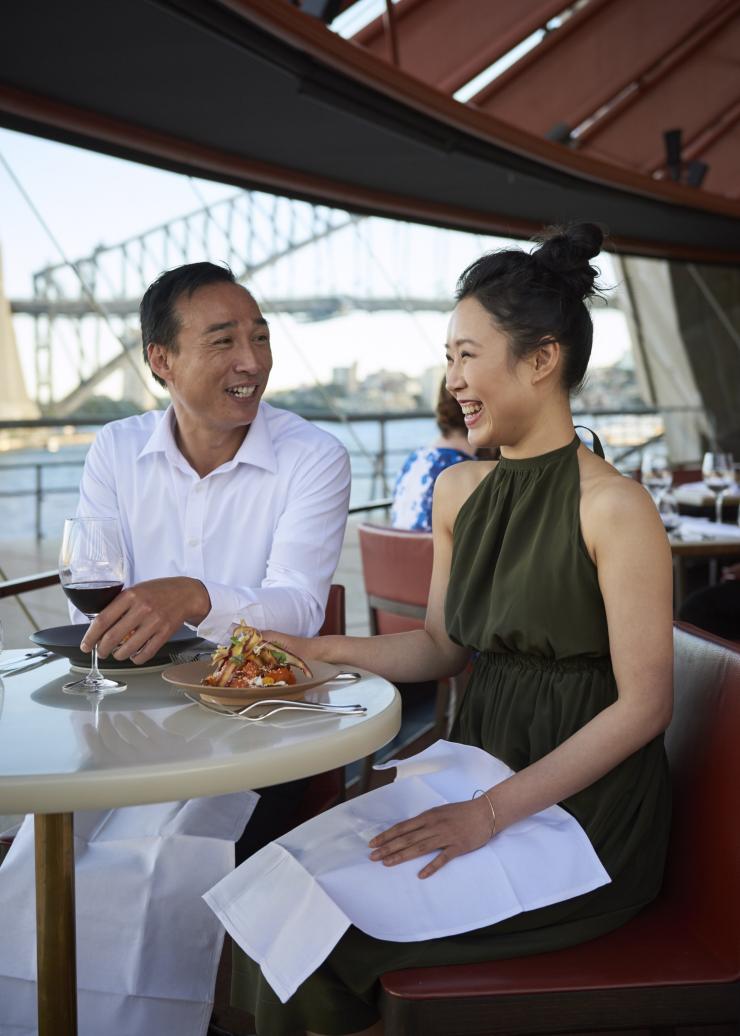 Un uomo e una donna seduti al ristorante Bennelong sorridono ammirando il Sydney Harbour Bridge sullo sfondo, Sydney, New South Wales © Destination NSW