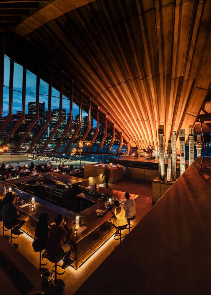 La sala dalle luci soffuse con ospiti seduti ai tavoli del ristorante Bennelong all'interno della Sydney Opera House, Sydney, New South Wales © Tourism Australia