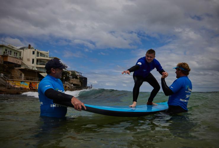 Uomo con disabilità neurologica che fa surf con istruttori del Let's Go Surfing, Bondi Beach, Sydney, New South Wales © Tourism Australia