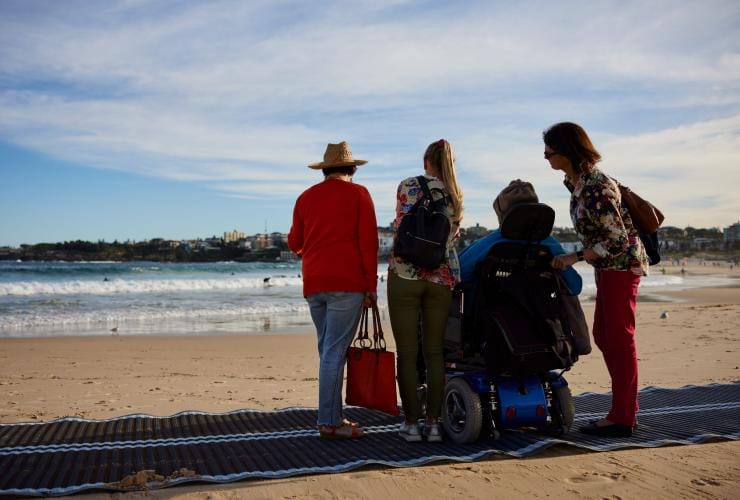 Uomo su una sedia a rotelle sulla spiaggia con la sua famiglia a Bondi Beach con Australia in Style, Sydney, New South Wales © Destination NSW