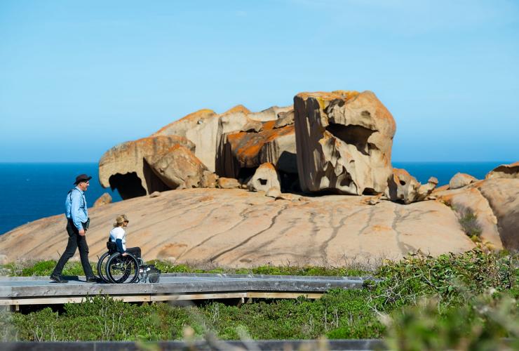 Una guida turistica che cammina con una donna su una sedia a rotelle accanto alle Remarkable Rocks, Kangaroo Island, South Australia © Tourism Australia