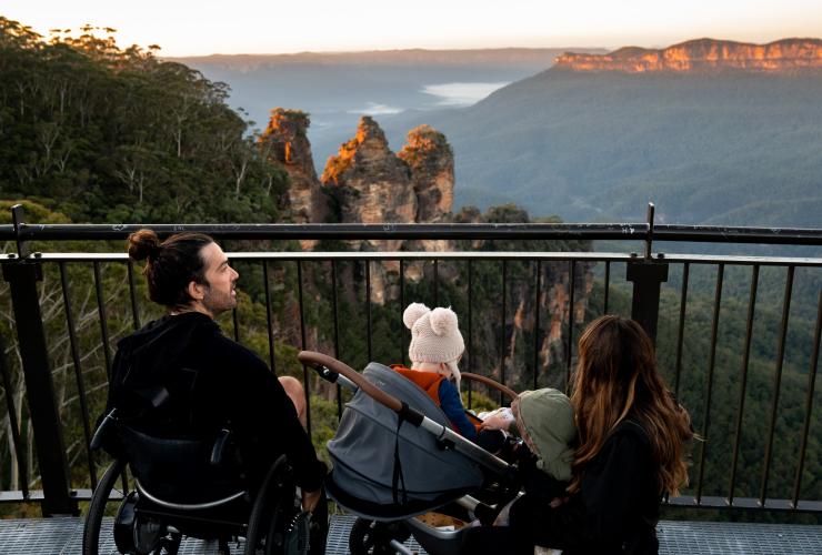 Uomo su una sedia a rotelle con la sua famiglia che ammirano le formazioni delle Three Sisters nelle Blue Mountains, New South Wales © Tourism Australia