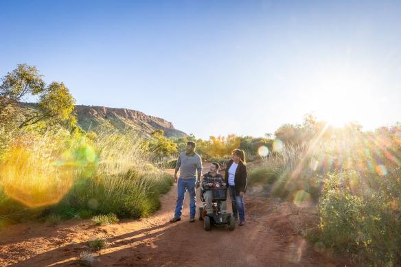Uomo con una disabilità neurologica su uno scooter elettrico con un altro uomo e una donna nell'Alice Springs Desert Park, Alice Springs, Northern Territory © Tourism NT/Helen Orr