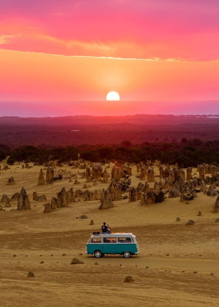 Due persone sedute sul tetto di un furgone che guardano verso alte formazioni rocciose sotto un tramonto rosato ai Pinnacles nel Nambung National Park, Coral Coast, Western Australia © Tourism Australia