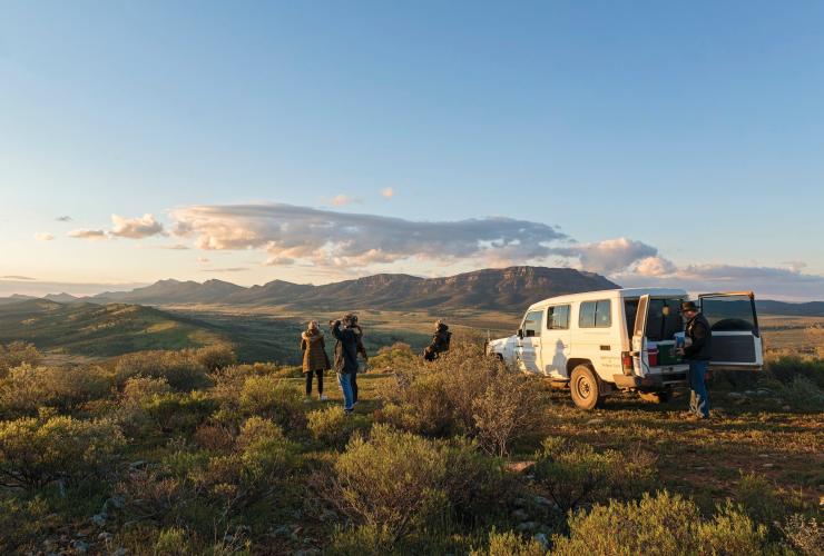 Un gruppo di persone accanto a un fuoristrada su una collina affacciata su un verde paesaggio dell'outback con montagne in lontananza alla Rawnsley Park Station, Flinders Ranges, South Australia © John Montesi