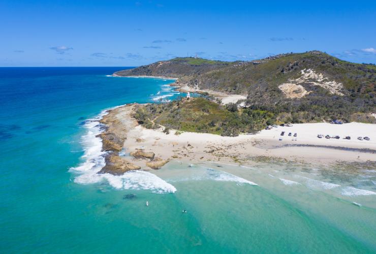 Vista dall'alto del promontorio di un'isola, con persone che fanno surf e nuotano in acque turchesi e una fila di auto parcheggiate sulla sabbia al North Point, Moreton Island, Queensland © Tourism and Events Queensland