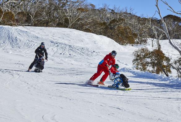 Due persone su sci per disabili che scendono con le loro guide da una montagna innevata al Thredbo Alpine Village, Snowy Mountains, New South Wales © Tourism Australia 