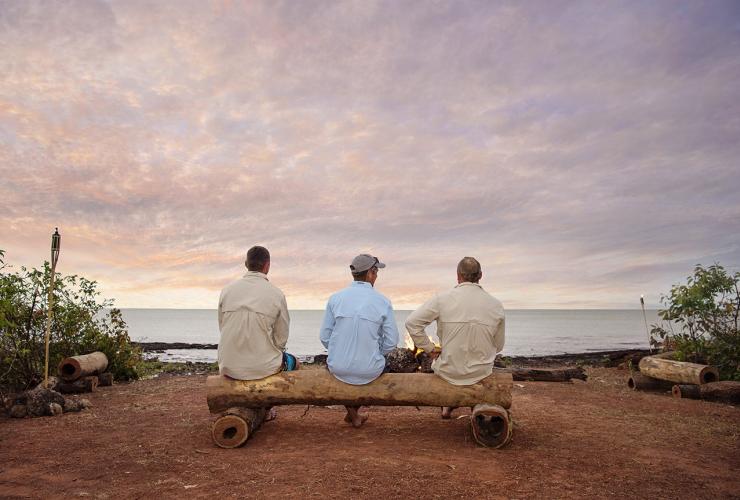 Tre persone sedute fianco a fianco su un tronco accanto a un falò in riva all’oceano, Dhipirri Barra & Sportfishing, Arnhem Land, Northern Territory © Shaana McNaught