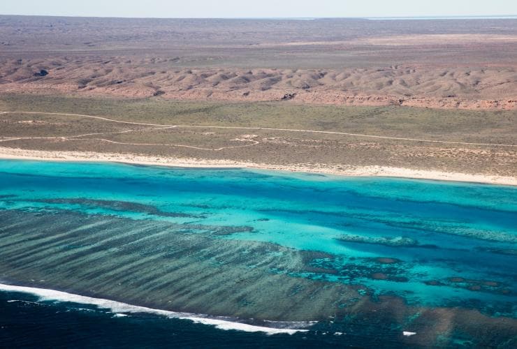 Veduta aerea di una costa di sabbia bianca con un paesaggio dell’outback in lontananza e un oceano cristallino con barriera corallina visibile, Ningaloo Reef, Western Australia © Tourism Western Australia