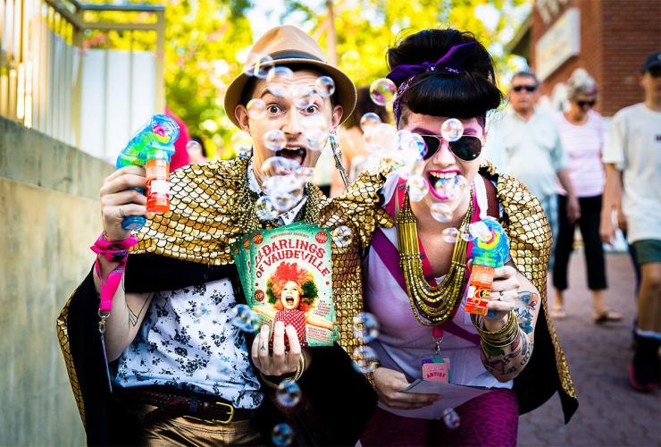 Due persone che indossano vestiti coloratissimi e soffiano bolle verso la fotocamera durante il Fringe World Festival, Perth, Western Australia © Artrage Inc