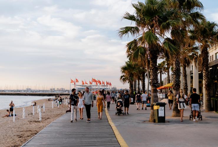 Gruppi di persone che passeggiano lungo il viale costeggiato dalle palme della spiaggia di St Kilda a Melbourne, Victoria © Visit Victoria