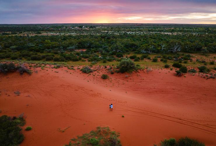 Vista dall'alto di madre e figlia in piedi su una vasta distesa di sabbia rossa con vista sul bushland al tramonto presso la Bullara Station, Ningaloo, Western Australia © Tourism Australia