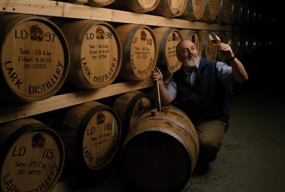Uomo accovacciato accanto a una parete di botti di rovere che ammira un bicchiere di liquore appena versato alla Lark Distillery, Hobart, Tasmania © Osborne Images