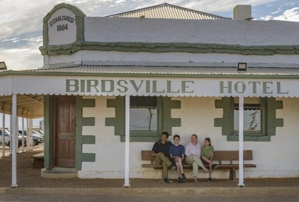 L'esterno di uno storico pub verde e bianco, con una famiglia di quattro persone sedure su una sedia sotto la veranda del locale al Birdsville Hotel, Queensland © Tourism and Events Queensland