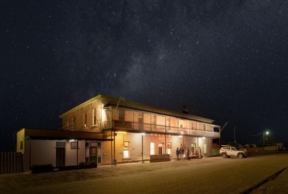 Persone in piedi in cerchio con delle bevande all'esterno di un locale di sera sotto le stelle al Marree Hotel, Flinders Ranges & Outback, South Australia © South Australian Tourism Commission